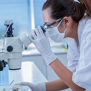 A women wearing a mask and gloves looking through a microscope - Serving Immigrants