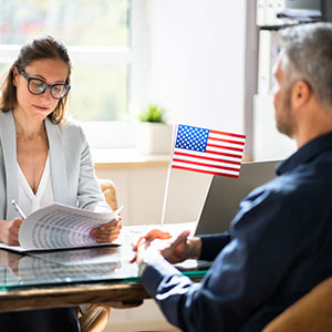 A woman and a man sitting at a table with an American flag in the background - Serving Immigrants